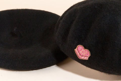 Close-up of black wool beret with sparkly pink crystal EMB cupcake pin detail on beige background