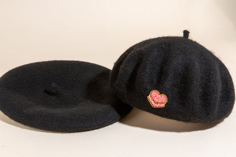 Two black wool berets from Erin McKenna's Bakery, one with removable pink crystal cupcake pin attached, styled on neutral background