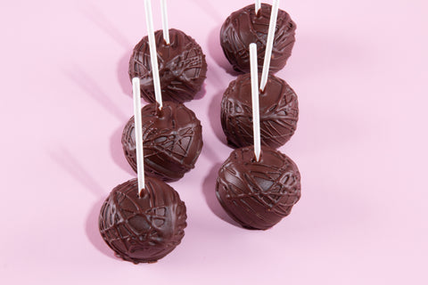 Six chocolate-coated cake pops on white sticks arranged on a soft pink background, each covered in dark chocolate with decorative drizzle patterns creating elegant crosshatch designs.