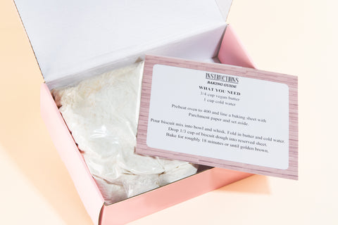 Open pink box revealing Erin McKenna's Bakery Butter Biscuit Mix contents including baking mix in clear bag and instruction card with baking guide against neutral background