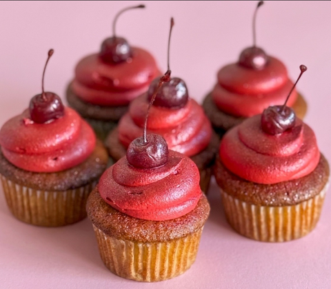 Six cupcakes with swirled red glitter frosting and a whole dark cherry on top, displayed on a glass cake stand in front of an ornate white plaster backdrop.
