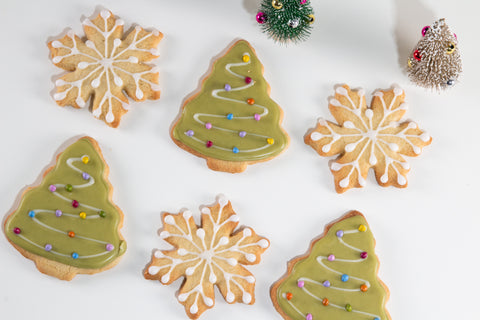 Overhead view of decorated holiday cookies on a white surface: three Christmas tree-shaped cookies with green matcha icing and colorful pearl sprinkles arranged in garland patterns, three snowflake-shaped cookies with white royal icing details, and two small bottle brush Christmas trees with metallic ornaments in the background.