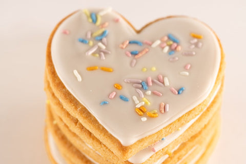A close-up of a stack of heart-shaped cookies, showing the top cookie with white icing and pastel-colored sprinkles. The cookies are arranged in a neat stack, highlighting their golden edges.