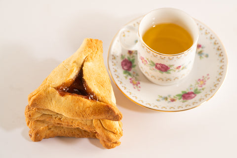 A stack of three golden-brown hamantaschen cookies filled with fruit jam sits next to a floral-patterned teacup on a matching saucer. The teacup is filled with light yellow tea, and both the cup and saucer feature pink and green rose designs with gold accents. The plain white background emphasizes the contrast between the delicate teacup and the cookies.