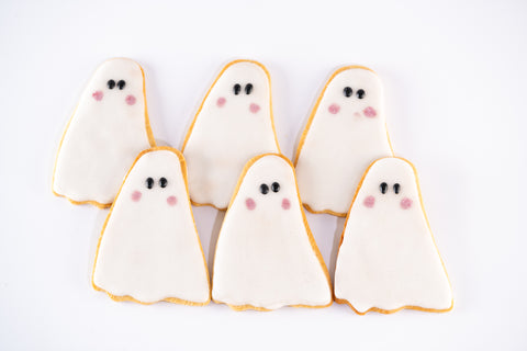 Ghost-shaped Halloween sugar cookies with white icing, black eyes, and pink cheeks, arranged on a white background.