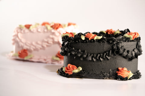 Side view of a coffin-shaped black mini cake with floral icing decorations and orange rose details, with a pink cake blurred in the background.