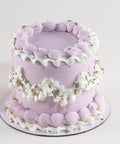 A round layer cake frosted in soft lavender, decorated with ridged lavender shell-border piping, ruffled white frosting garlands, and scattered dried lavender buds, displayed on a lavender cake board against a white background.