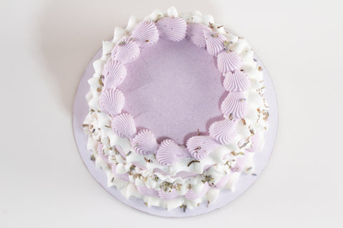 Overhead view of a round lavender cake with a ring of ridged lavender shell-piped frosting and ruffled white frosting accented with dried lavender buds, on a lavender cake board.