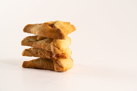 A stack of four golden brown, triangular-shaped pastries neatly arranged on a white surface. The flaky dough appears soft and slightly textured, with a hint of fruit filling peeking out between layers. The pastries are photographed against a plain, light background, emphasizing their rich, baked texture and the subtle color variations in the crust.