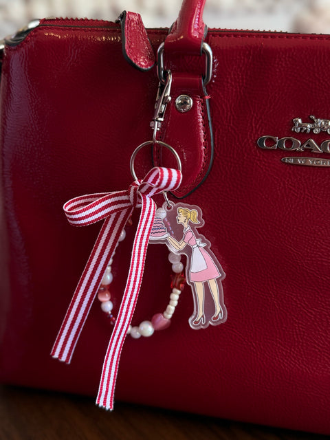 A red patent leather Coach handbag with a decorative bag charm clipped to the handle. The charm features a red and white candy-stripe grosgrain ribbon bow, a loop of pink, pearl, and red glass beads, and an acrylic charm of a retro illustrated woman in a pink dress and heels holding a tiered cake.