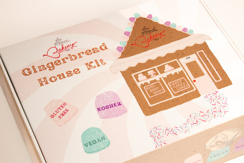 Erin McKenna's Bakery Gingerbread House Kit packaging featuring bakery logo, brown gingerbread house illustration, and three heart-shaped badges indicating 'Gluten Free', 'Kosher', and 'Vegan' certifications. The house illustration includes a decorated storefront with 'Baby Baby Baby' and 'Cakes Cakes Cakes' text