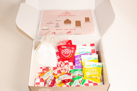 Open gingerbread house kit box showing instruction sheet labeled 'What's Inside?' and 'What You Need!' with illustrations of required supplies. Below are various colorful YumEarth candy packages and giggles candy on red and white checkered paper