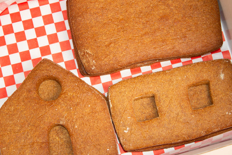 Close-up of baked gingerbread house pieces on red and white checkered paper, showing golden brown cookie walls with cut-out windows and doors, displaying the characteristic texture of gingerbread