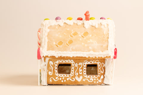 Overhead view of unassembled gingerbread house pieces arranged in a white box on red and white checkered paper, showing the front wall with circular windows and side walls with square cut-outs