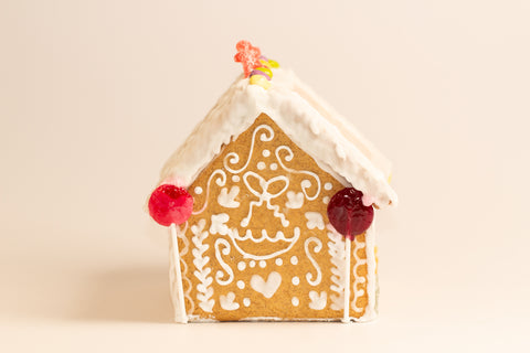 Decorated gingerbread house front view showing white frosting details around windows, decorative piped patterns, and colorful candies along the roofline including yellow, purple, and red gummy candies