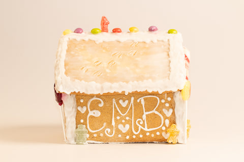 Front view of gingerbread house decorated with 'EMB' in white icing surrounded by hearts and dots, white icing roof trim, colorful candies along the roofline, and small gummy figures at the base