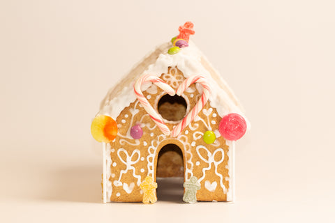 Close-up front view of gingerbread house showing detailed white icing decorations of gingerbread people and hearts, a candy cane heart above the doorway, colorful round candies on the sides, and gummy bear decorations at the bas