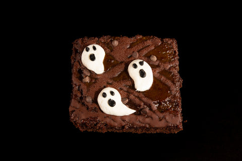 Gluten-free vegan chocolate brownie with three white frosting ghosts and chocolate chips on black background - Halloween ghost brownie top view