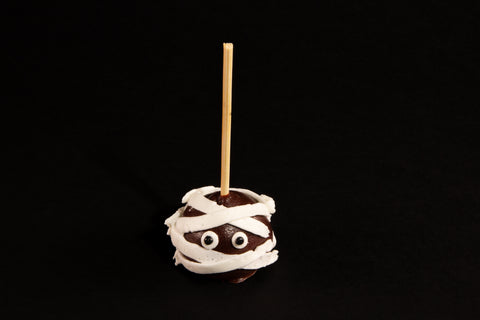 Chocolate mummy cake pop with white frosting bandages and candy eyes on wooden stick against black background - gluten-free vegan Halloween treat