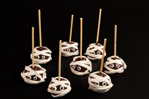 Group of eight chocolate mummy cake pops decorated with white frosting wraps and googly eyes on wooden sticks - allergy-friendly Halloween desserts