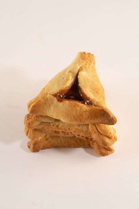 A stack of three golden-brown hamantaschen cookies with slightly crimped edges. The top cookie has a visible filling of sweet fruit jam in the center, with the three corners folded over to form a triangular shape. The background is plain and white, emphasizing the texture and color of the cookies.