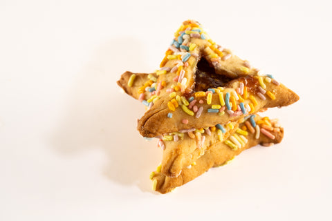 Two stacked hamantaschen cookies decorated with colorful sprinkles. The cookies are triangular in shape, with a visible fruit jam filling peeking out from the top. The edges of the cookies are covered in yellow, blue, pink, and white sprinkles, adding a festive appearance. The background is plain white, highlighting the bright sprinkles and the cookie's golden-brown color.