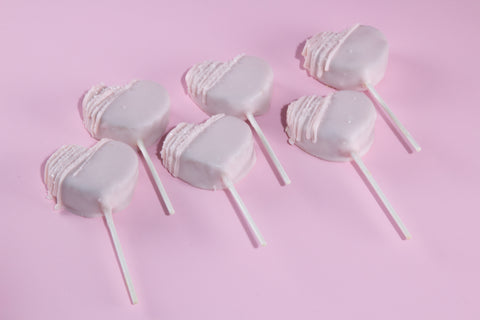 Heart-shaped vegan cake pops with white chocolate coating and pink drizzle on pink background.