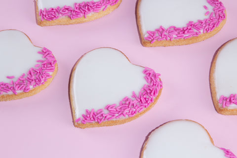 Heart-shaped sugar cookies decorated with white royal icing and pink sprinkles arranged on pink background
