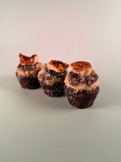 Three miniature chocolate cupcakes with sugar-coated tops and caramel drizzle, arranged on a neutral light gray background.