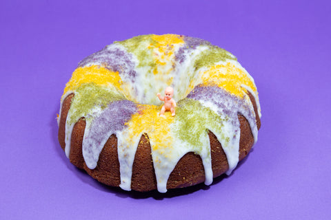 Celebratory vegan and kosher King Cake featuring traditional Mardi Gras decorations with baby figure nestled among colorful sugar crystals on bright purple background.