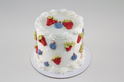 A white-frosted layer cake decorated with hand-sculpted strawberry, blueberry, and raspberry decorations with green leaves, finished with piped rosette borders, on a white cake board.