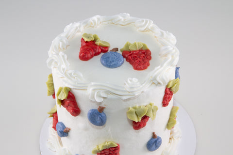 Close-up top view of a white layer cake with hand-sculpted berry decorations including strawberries, blueberries, and raspberries with green leaves, and a piped rosette border.