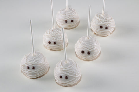 Six Halloween-themed cake pops decorated to look like wrapped mummies, with white frosting, white lollipop sticks, and small brown candy eyes, arranged on a light gray background.