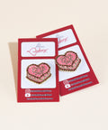Two EMB 2025 enamel pins featuring pink cake design with chocolate frosting detail and vintage gold lettering on red branded backing cards from Erin McKenna's Bakery
