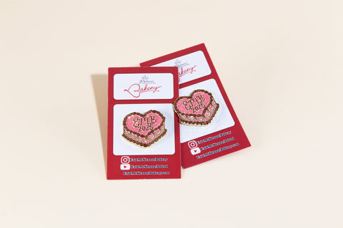 Two EMB 2025 enamel pins featuring pink cake design with chocolate frosting detail and vintage gold lettering on red branded backing cards from Erin McKenna's Bakery
