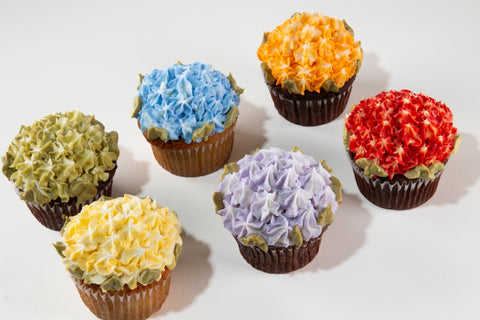 Six cupcakes with densely piped floral frosting in Pride rainbow colors — green, blue, orange, yellow, purple, and red — with sage green leaf accents, arranged on a white surface.