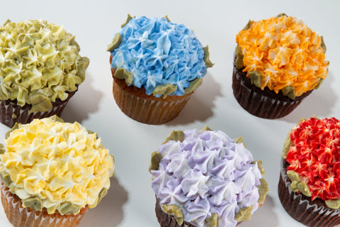 Overhead view of six cupcakes in a 2x3 grid, each topped with densely piped floral frosting in a different Pride rainbow color — green, blue, orange, yellow, purple, and red — with sage leaf accents.