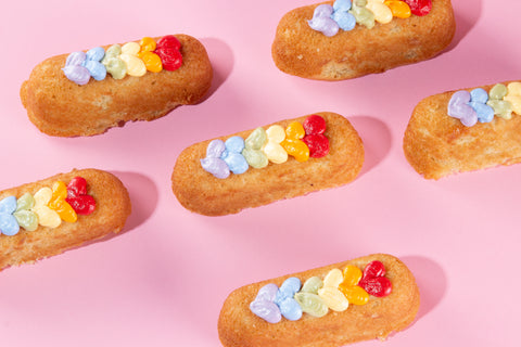 Six golden sponge cake fingers scattered on a pink background, each topped with a row of small rainbow-colored heart-shaped frosting dots in Pride flag colors.