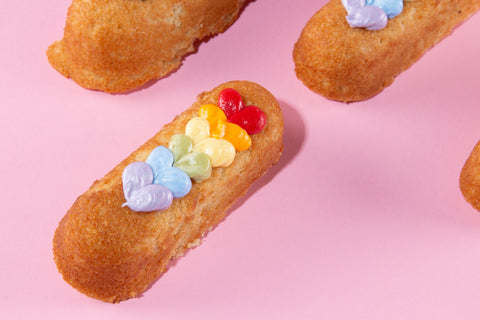 Close-up of a single golden sponge cake with a diagonal row of rainbow frosting hearts in purple, blue, green, yellow, orange, and red on top, on a pink background.