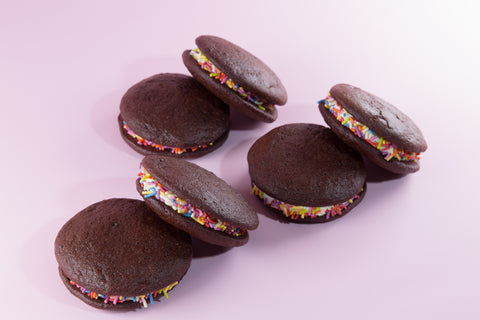 Six chocolate whoopie pies arranged in a loose cluster on a pink background, each showing a thick cream filling studded with colorful rainbow sprinkles along the edges.