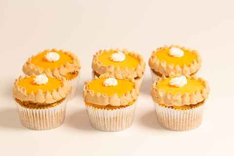 Six vegan gluten-free pumpkin pie cupcakes arranged in two rows, each topped with smooth orange pumpkin filling, tan piped frosting borders, and white whipped cream centers