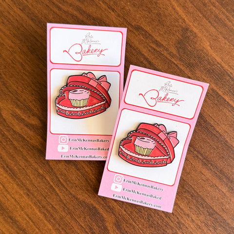 Two cupcake enamel pins featuring pink cupcake design in a red, chocolate candy box