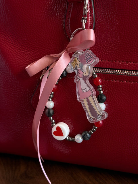 A red patent leather handbag with a decorative bag charm clipped to the zipper pull. The charm features a dusty pink satin ribbon bow, a beaded loop mixing black marble, red, silver, and white pearl beads with a white heart charm, and an acrylic charm of a retro illustrated woman in a pink apron dress holding a dark layered cake, with "EMB" printed on her apron.