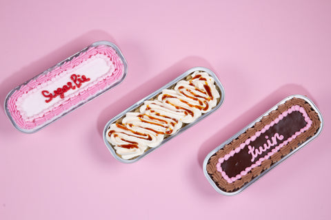 Three mini tinned cakes arranged on pink background: pink-frosted vanilla, caramel-drizzled tres leches, and chocolate raspberry with decorative piping