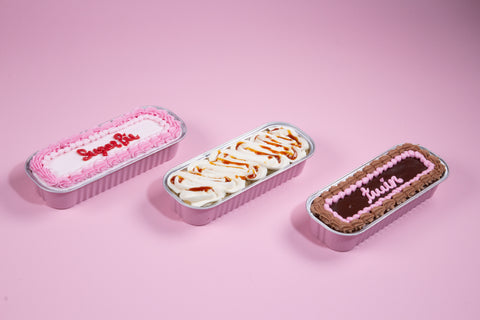 Three mini tinned cakes arranged on pink background: pink-frosted vanilla, caramel-drizzled tres leches, and chocolate raspberry with decorative piping