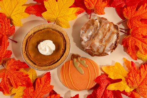 Overhead view of vegan Thanksgiving dessert trio featuring 3-inch pumpkin pie topped with whipped cream, glazed pumpkin bundt cake, and festive pumpkin-shaped cookie surrounded by red, orange, and yellow fall foliage