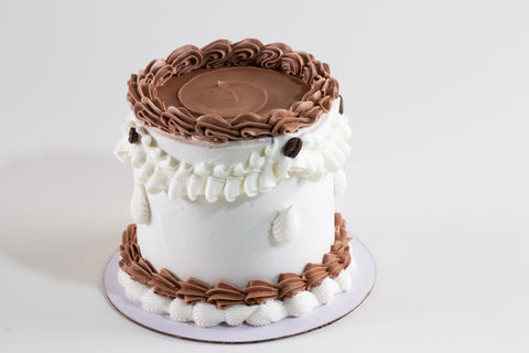 A round cake with white frosting and brown piped borders, topped with a smooth brown layer and decorated with white shell-shaped frosting details, shown on a white background.