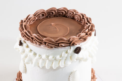 A close-up of a decorated cake with a smooth chocolate brown top, piped chocolate rosettes around the edge, white frosting on the sides, and a few dark chocolate chips.