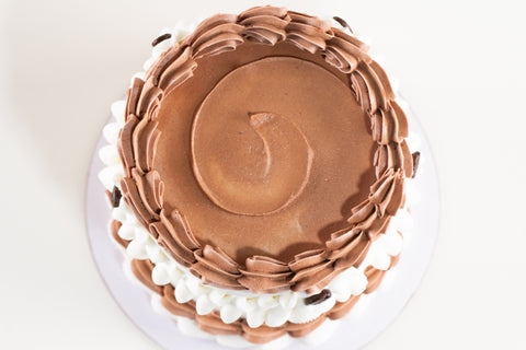 A round chocolate cake with piped brown frosting on top and sides, decorated with white frosting borders, shown from a top-down view on a white plate against a white background.