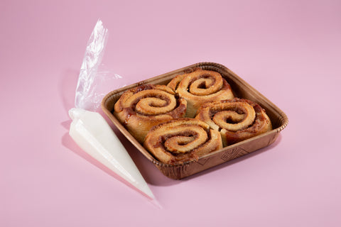 Four golden-brown cinnamon rolls in a brown paper baking tray with a piping bag of white frosting on a soft pink background.